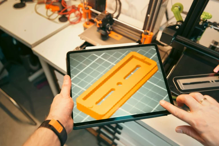 Eine Person zeigt eine orangefarbene 3D-gedruckte Komponente auf dem Tablet vor einem 3D-Drucker