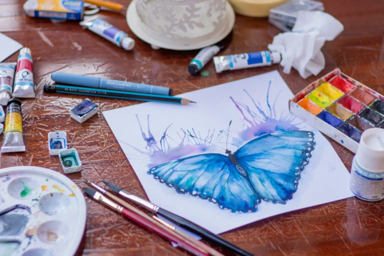 Aquarellzeichnung eines blauen Schmetterlings auf Papier, umgeben von Malutensilien auf einem Holztisch