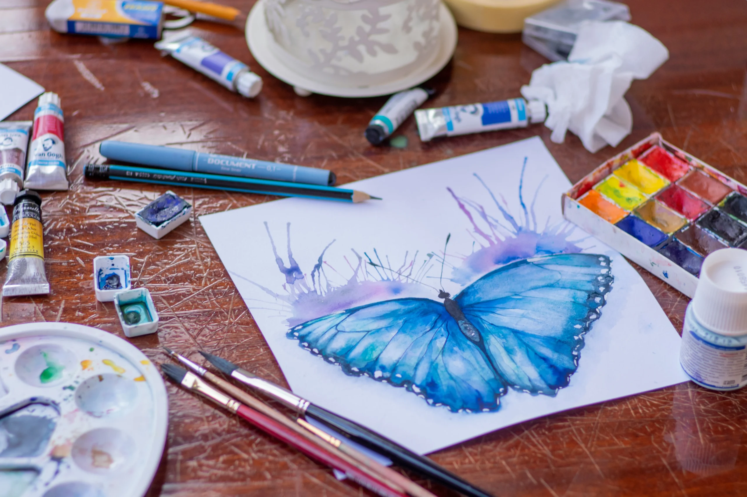 Aquarellzeichnung eines blauen Schmetterlings auf Papier, umgeben von Malutensilien auf einem Holztisch